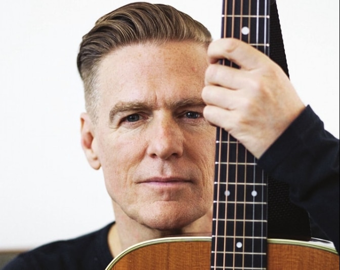A portrait shot of Bryan Adams staring at the camera holding his guitar in a studio