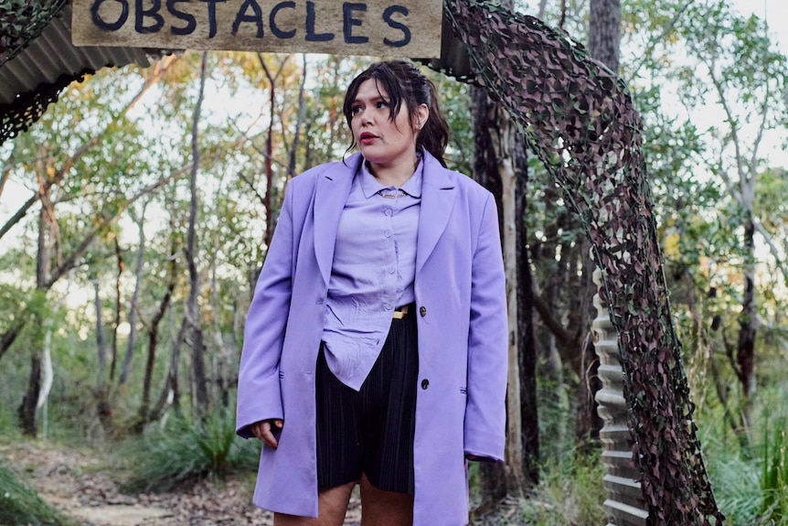 An Aboriginal woman wearing purple stands in the bush under a sign: "obstacles".