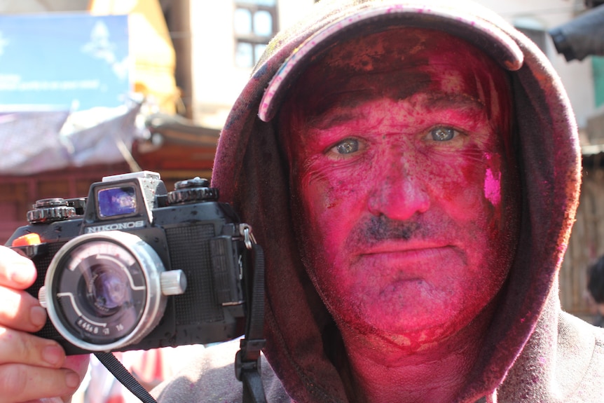 Man with bright pink powder on face holding camera up to the side of his head.