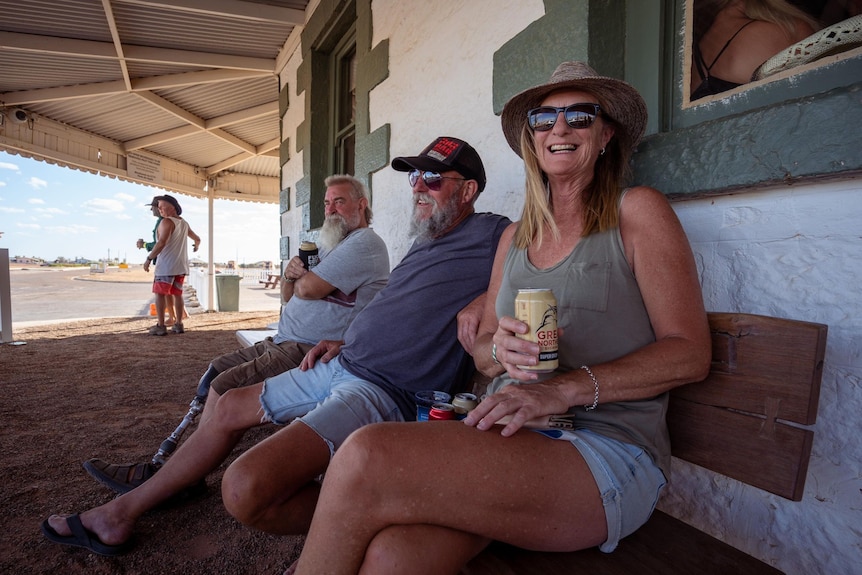 A smiling woman with denim shorts, grey singlet, holding a beer, sits on a wooden bench under a verandah with two other men.