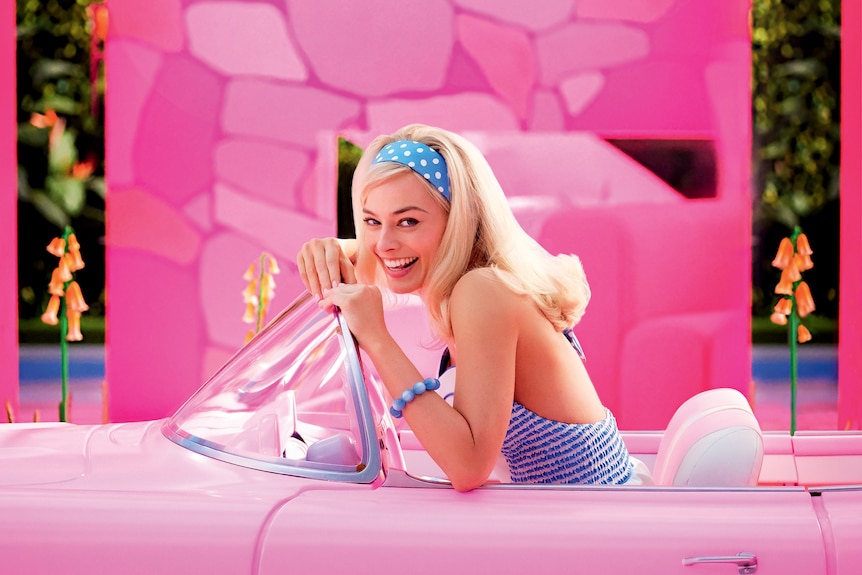 A blonde white woman wearing a blue and white striped halter-neck and matching headband drives a bright pink car.