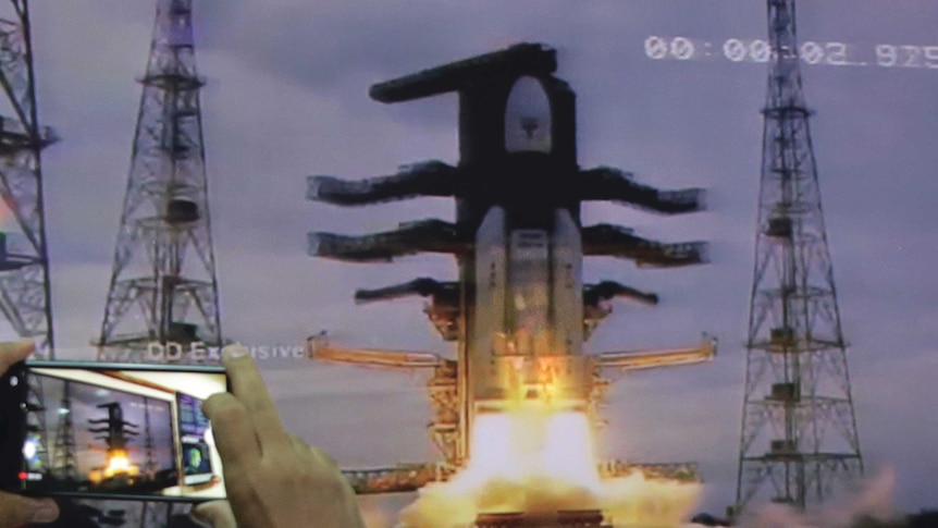 A rocket takes off with fire underneath it against a stand. A hand is seen on the left hand side of the image holding a phone.