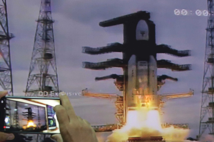 A rocket takes off with fire underneath it against a stand. A hand is seen on the left hand side of the image holding a phone.