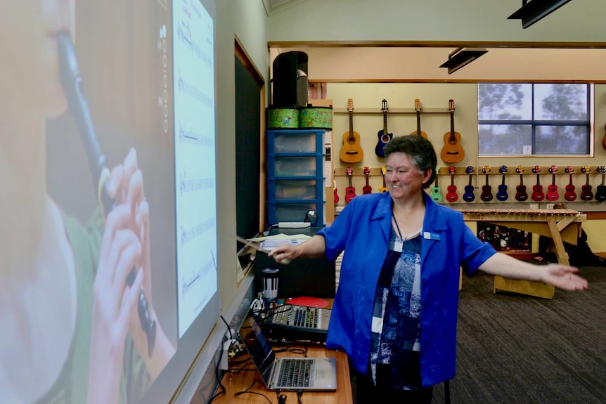 A woman points at a sheet of music projected onto a whiteboard in a music classroom.