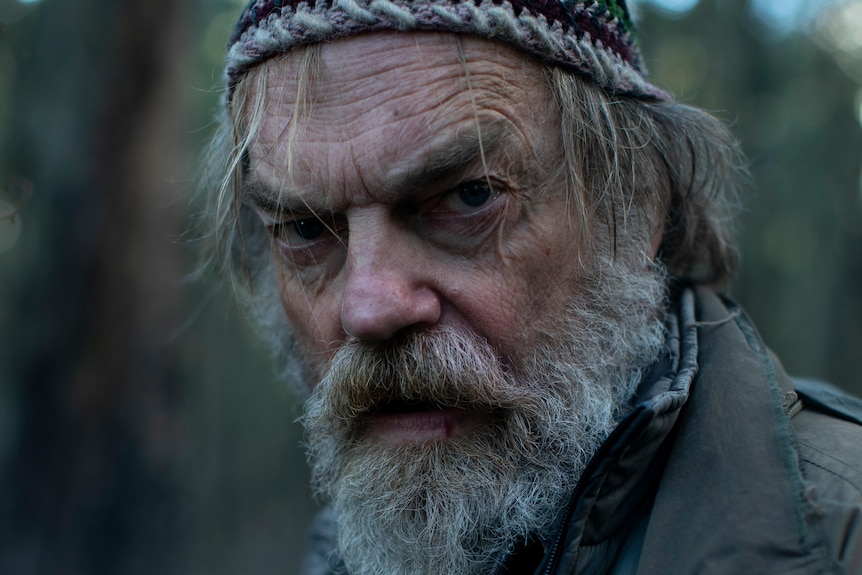 A film still of Hugo Weaving. He is wearing a beanie, has a full beard, and looks disheveled.