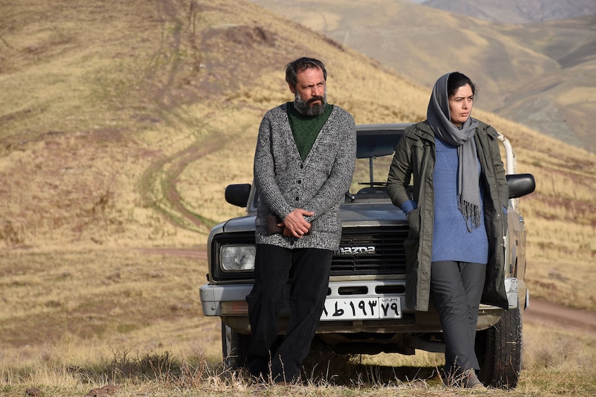 A 50-something man and 40-something woman lean against the bonnet of a truck, with dry grassy hills behind them.