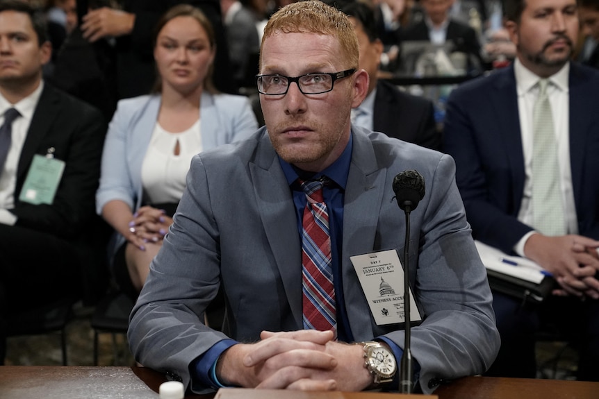 Мужчина в сером костюме и красно-синем клетчатом галстуке сидит у микрофона, сложив руки на столе.