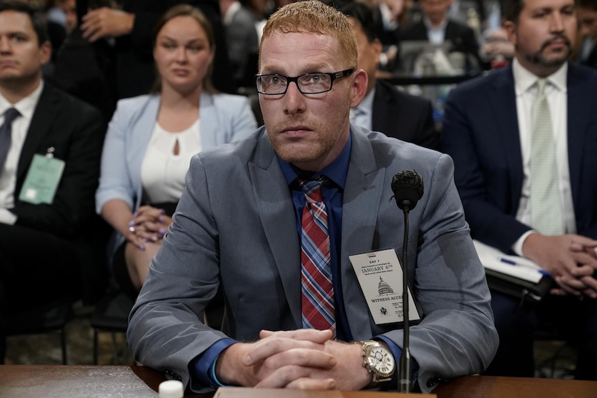 A man in a grey suit and red-and-blue-checked tie sits at a microphone with hands folded on the desk