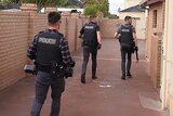 Three police officers in plain clothes and bullet-proof vests walk down the driveway of a property with objects in hand.