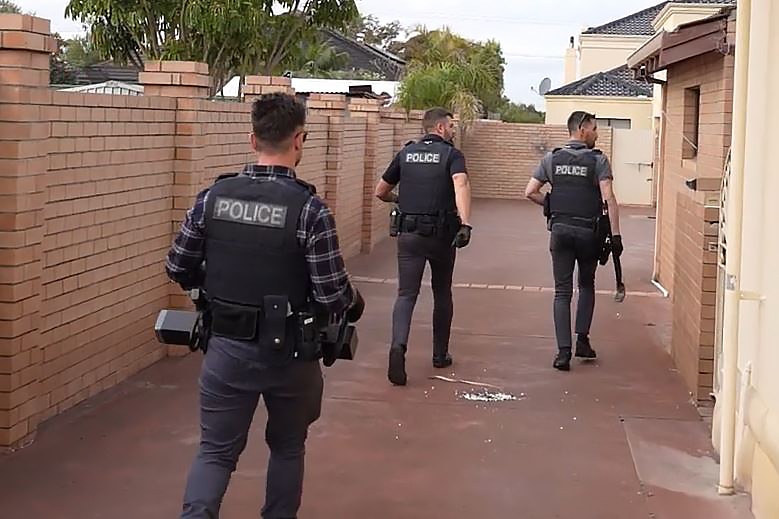 Three police officers in plain clothes and bullet-proof vests walk down the driveway of a property with objects in hand.