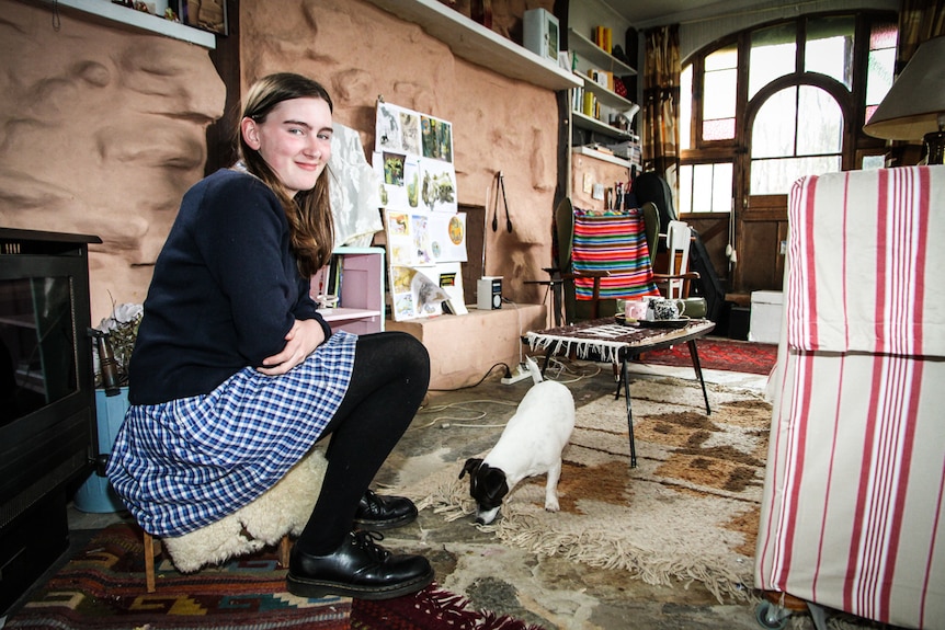 A girl in school uniform sits by a wood heater, with a small dog at her feet.