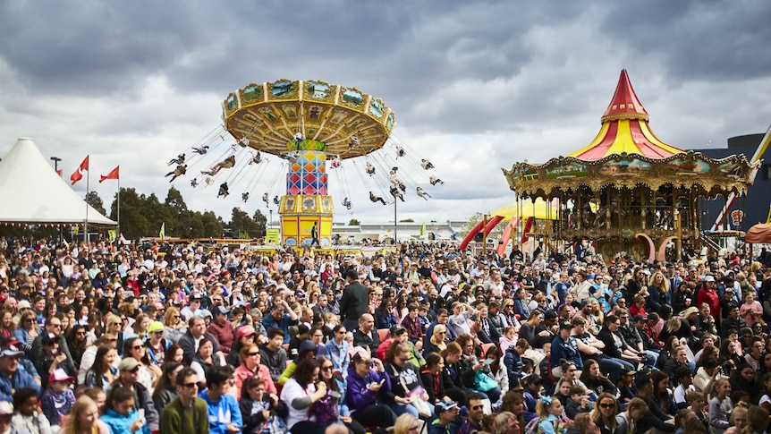 A large crowd gathers under a grey sky. There are rides and marquees in the background.