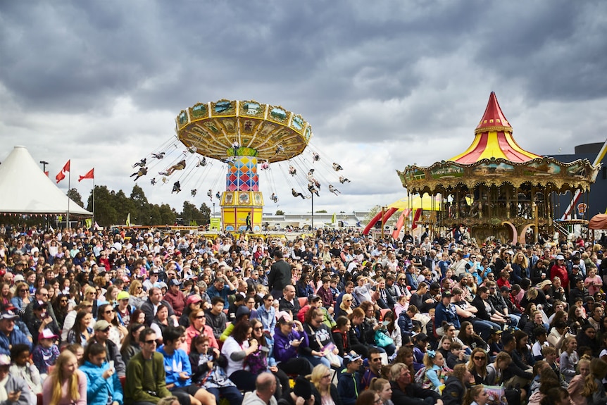 A large crowd gathers under a grey sky. There are rides and marquees in the background.