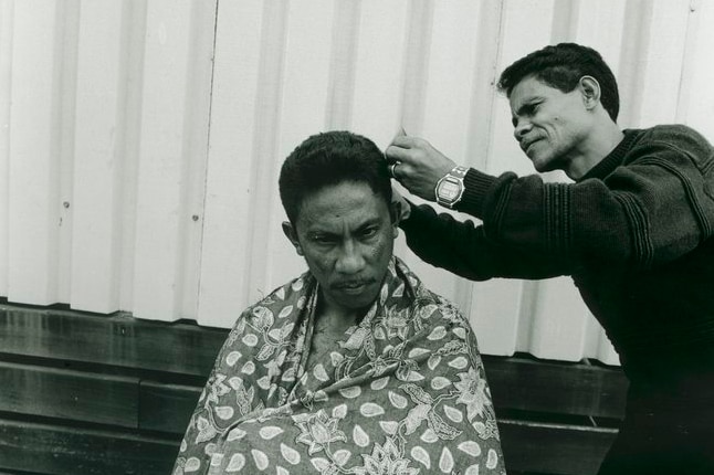 A man seated outside a prefabricated building in Puckapunyal, while another East Timorese man cuts his hair, in 1999.