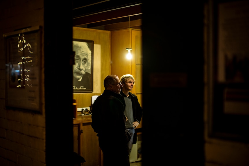 An older and young man stand in a room in front of Albert Einstein framed poster under a hanging warm hued lightbulb.