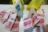 A person's gloved hands sprays disinfectant on Chinese banknotes. The person is wearing a yellow plastic coat.