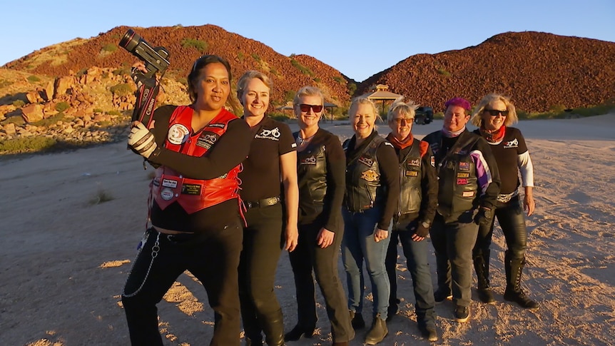 A group of women pose for a photo in the sunset beside a motorbike.