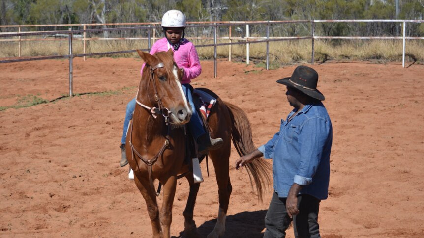 A man stands next to a horse, as a young Aboriginal girl sits on the saddle.