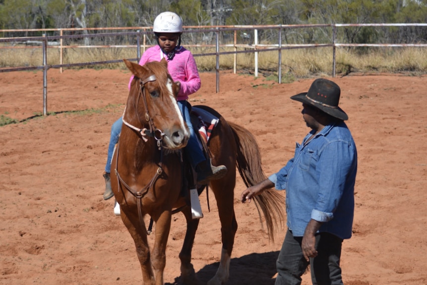 A man stands next to a horse, as a young Aboriginal girl sits on the saddle.