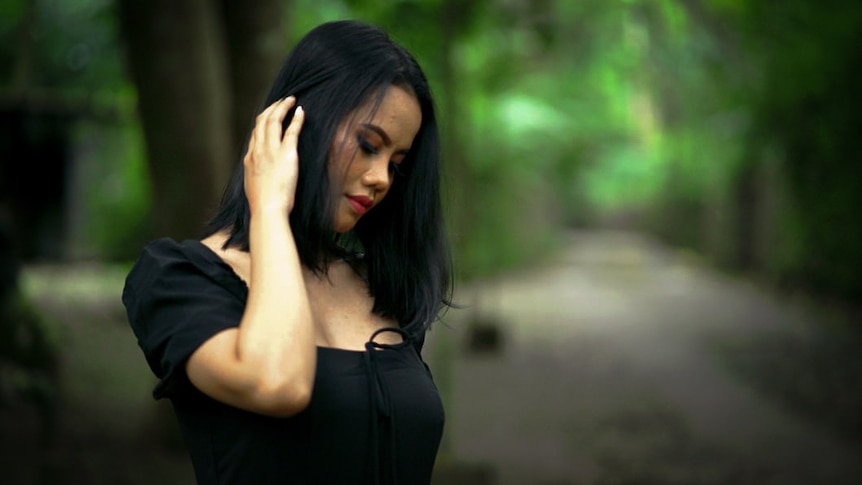 A woman wearing a black shirt tucks her hair behind one ear as she stands in the middle of a forest path