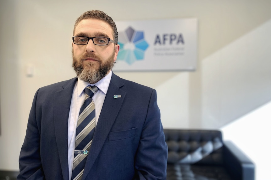 A man wearing a suit, with a beard and glasses looks at the camera 