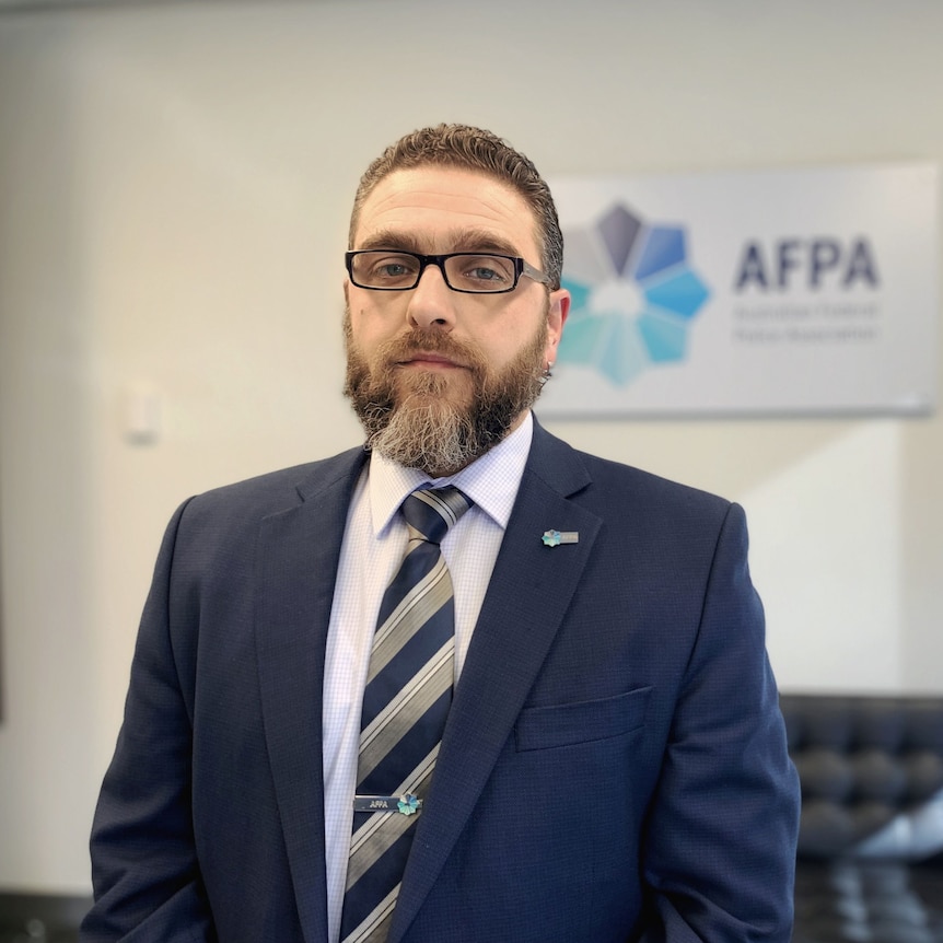 A man wearing a suit, with a beard and glasses looks at the camera 