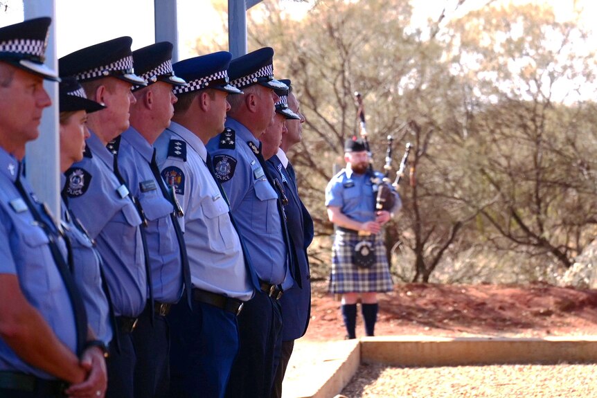 A line of police officers stand with a police bagpiper in the background