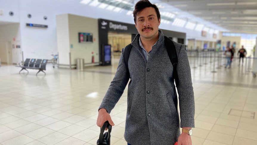 Justin Holland in grey jacket and holding his boarding pass, stands with his bag at the airport