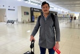 Justin Holland in grey jacket and holding his boarding pass, stands with his bag at the airport