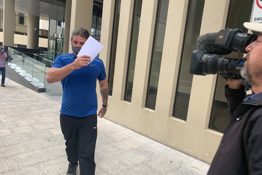 A man in a blue shirt walking out of court, shielding his face with a piece of paper.