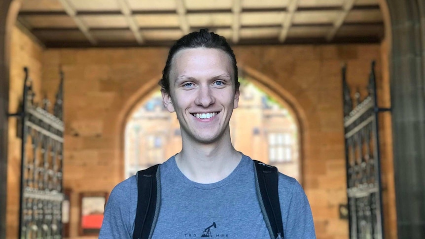 A young man smiles in front of a grand university sandstone archway.