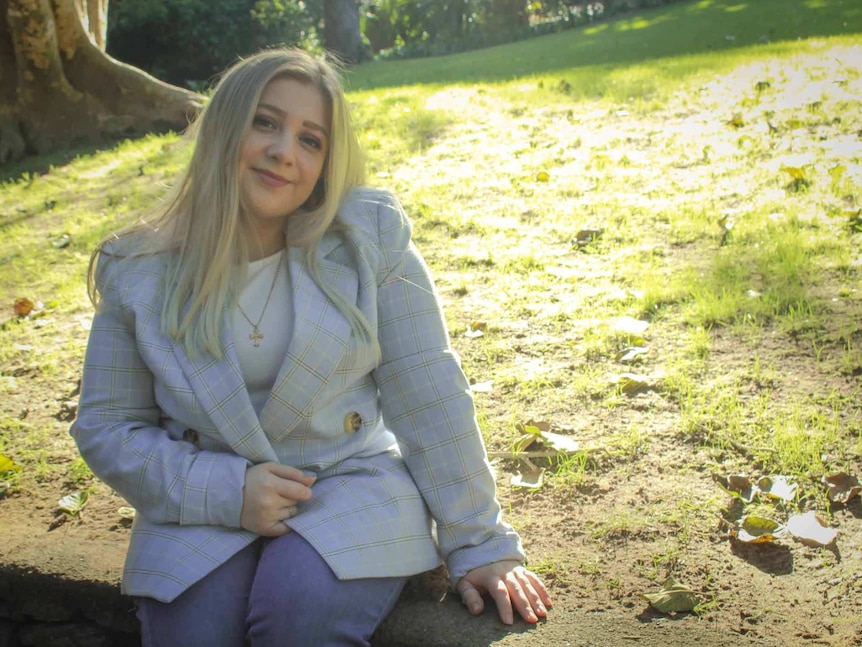 A smiling blonde woman in a grey suit jacket sits on a retaining wall on the edge of a grassy slope.