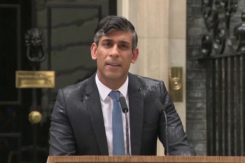 A man in a wet suit speaks into microphones at a lectern.