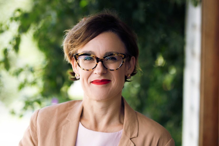 A woman with glasses smiles as she leans against a table top.