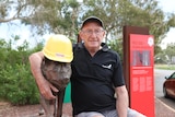 A man sits with a statue of a dog, which has a hard hat on its head with a sticker saying, 'visitor'.