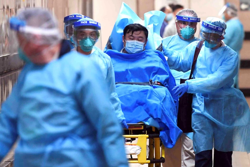 A man on a gurney wears a mask as doctors in larger masks push him through a hospital corridor.