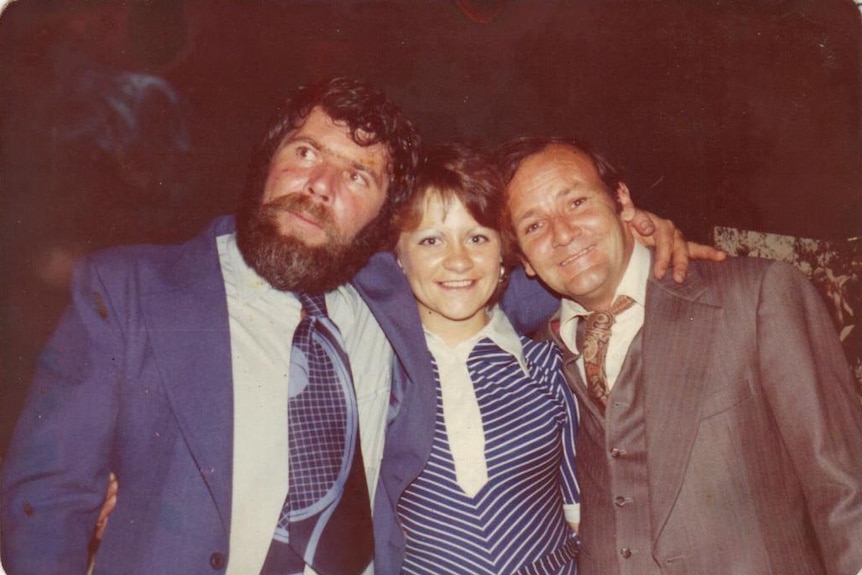 Two men in suits on either side of a woman in a striped dress smile at the camera 