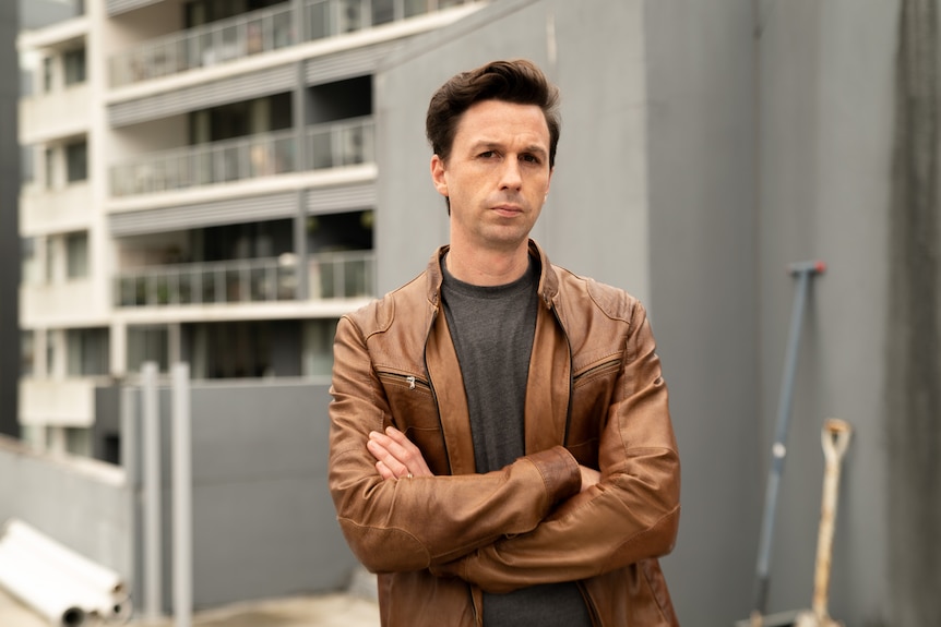 A man wearing a brown leather jacket, with his arms crossed, looks seriously at the camera with apartments in background