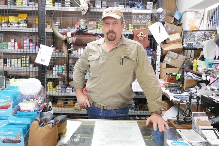 A man in a khaki shirt and cap stands behind a counter in a shop.