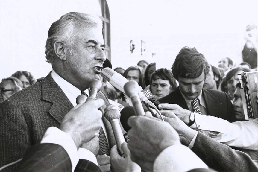 A man standing on the Parliament House steps surrounded by people and microphones.