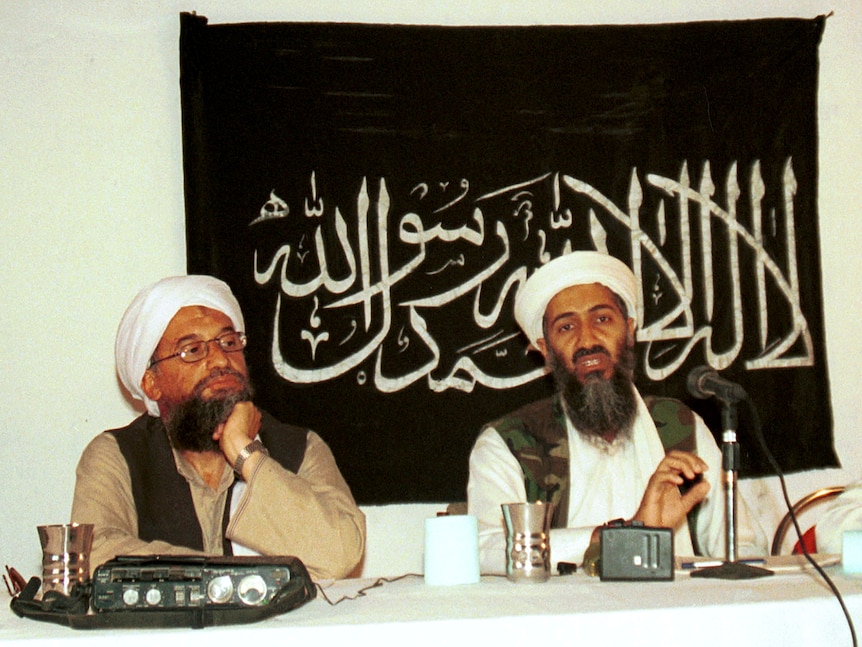 Two bearded men, including Osama Bin Laden, wearing turbans and waistcoats sit in front of a black flag bearing white writing