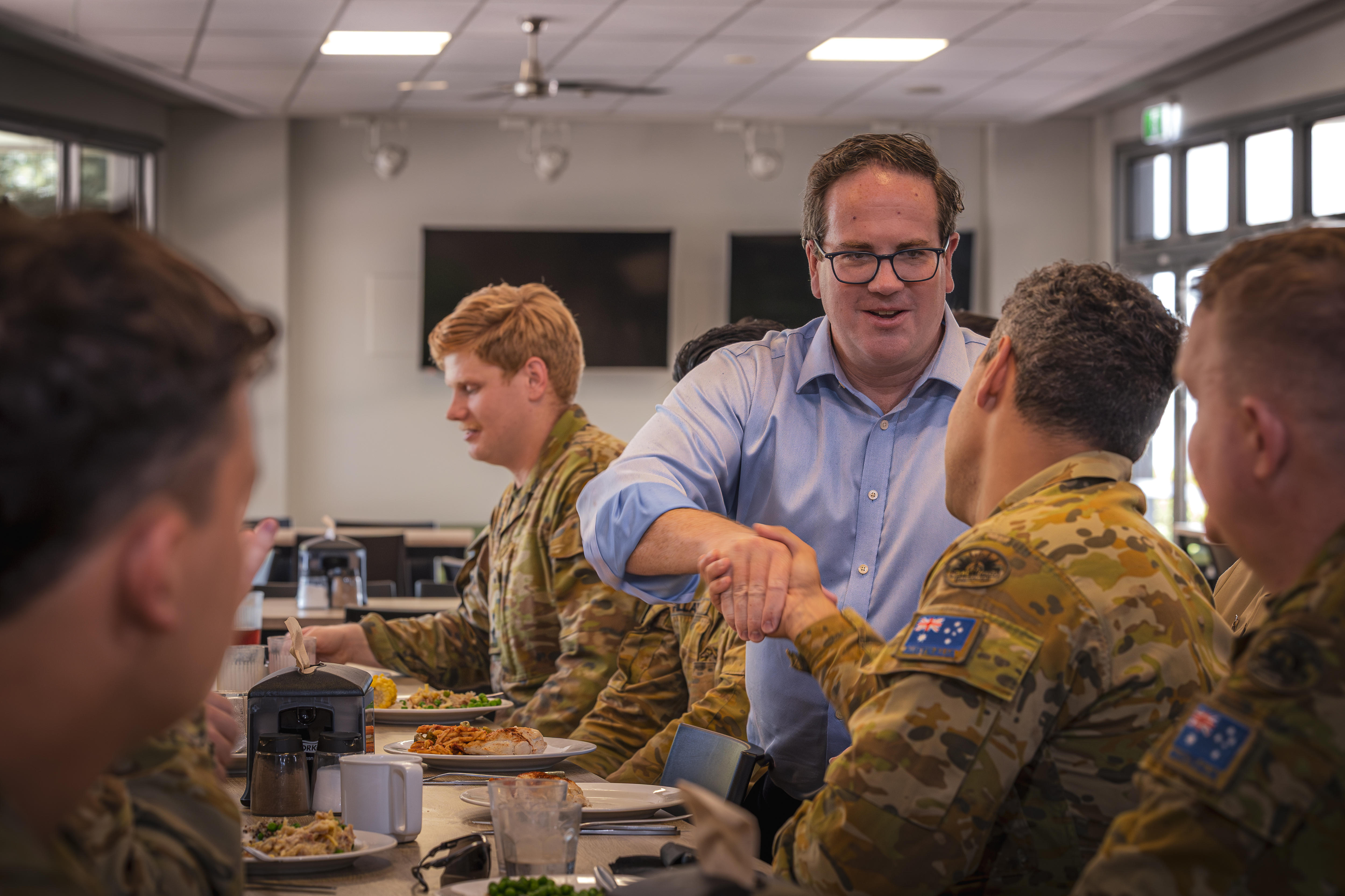Um homem de óculos fica entre homens em uniforme militar e aperta a mão de um deles.