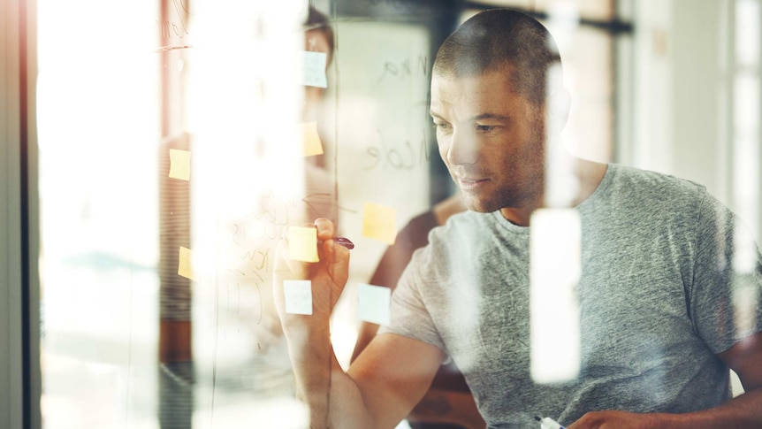 Man in t-shirt writing on post-it notes stuck on a glass wall