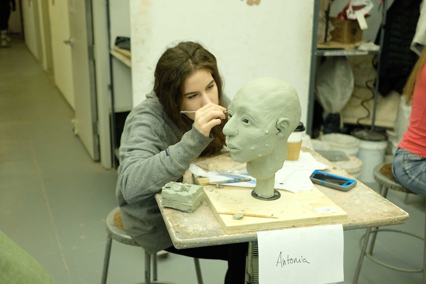 A young art student works on a sculpture of a human face.