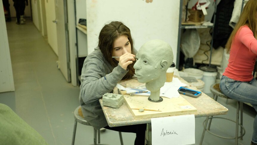 A young art student works on a sculpture of a human face.
