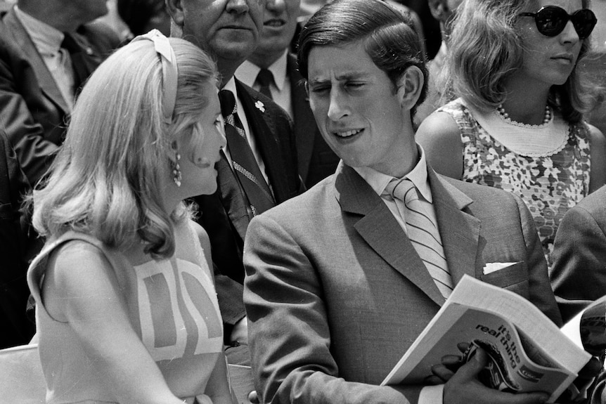 Prince Charles wearing a suit and reading a newspaper sits and talks to Tricia Nixon