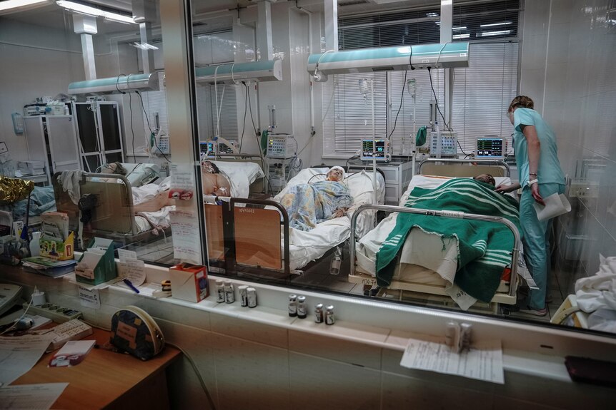View through a window to an intensive care ward with four patients in their beds and a nurse checking on the one furthest right.