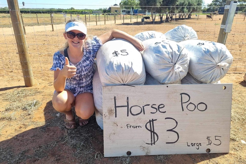 Girl on farm crouches down next to bags of horse manure, a sign says the bags are three dollars each.