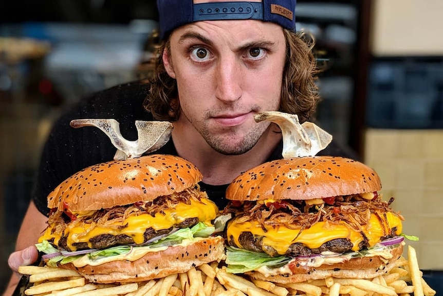A man in a backwards baseball cap poses with two enormous burgers on a mountain of fries.