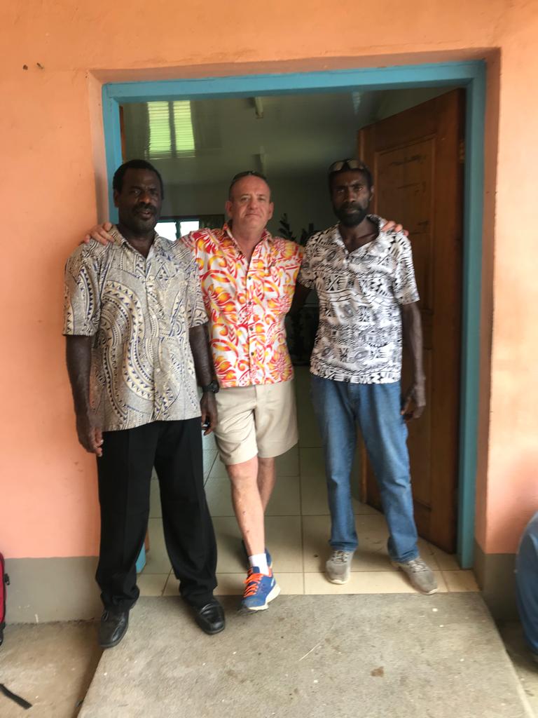 Two ni-Vanuatu men wearing pants and island-themed shirts with an Australian man in the middle wearing an orange floral shirt an